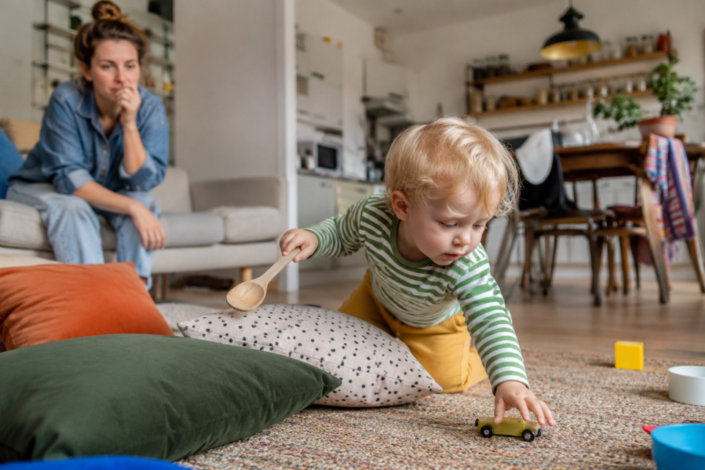Kleinkind spielt im Wohnzimmer mit Alltagsgegenständen, während seine Mutter zusieht